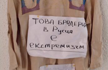 Ленена риза с бродерия «Молитва» е окачена на закачалка на текстурирана бяла стена. Към гърба на ризата с кабари е закрепен бял лист хартия А4 с ръкописен текст на български език: «ТОВА БРОДЕРИЯ В Русия е екстремизъм». Отдолу има по-малък лист с по-дълъг ръкописен текст